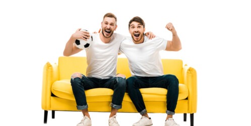 Excited,young,man,sitting,on,couch,with,soccer,ball,isolated